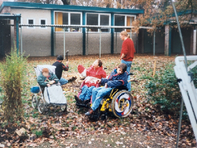Gartenansicht. Drei Kinder in Rollstühlen, dahinter ein sitzendes Kind und ein stehender Erwachsener.