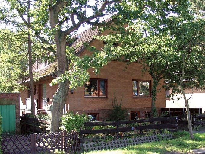 Hausfassade mit zwei Fenstern, umgeben von Bäumen und Garten