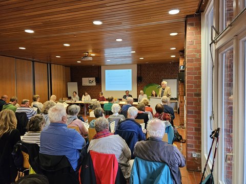 Mitgliederversammlung: Podium mit Menschen und Publikum auf Stühlen. 