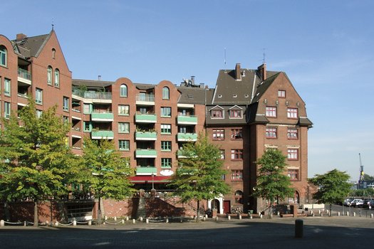 Außenansicht des Wohngebäudes Am Fischmarkt. Mehrstöckiger Klinkerbau mit grünen Balkonen. Davor Bäume und ein leerer Platz. 