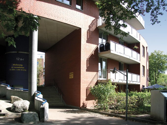 Eingang eines mehrstöckigen Wohngebäudes mit Balkonen und Treppe, umgeben von Bäumen und Sträuchern