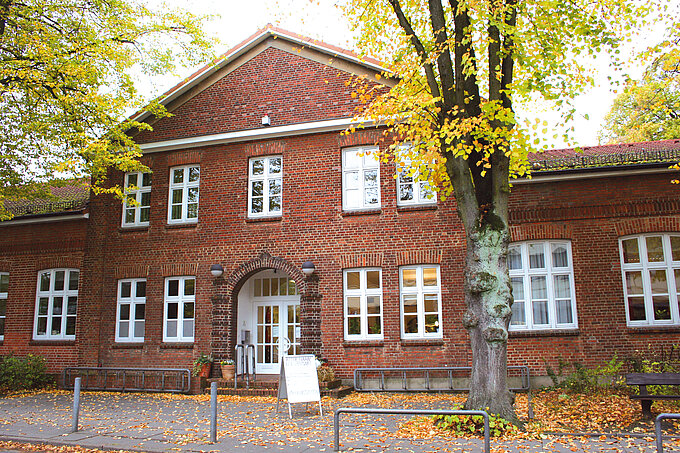 Backsteingebäude mit weißen Fenstern und Eingangstür, umgeben von Bäumen mit herbstlichem Laub