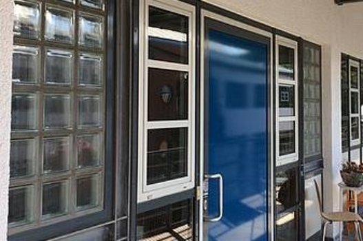 Eingang eines Gebäudes mit blauer Tür und Fenstern, darüber ein rotes rundes Schild mit weißer Schrift
