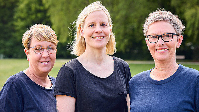 Drei Frauen stehen draußen auf einer Wiese, im Hintergrund Bäume