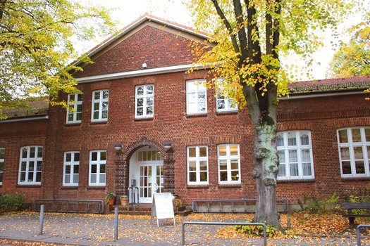 Backsteingebäude mit weißen Fenstern und Eingangstür, umgeben von Bäumen mit herbstlichem Laub