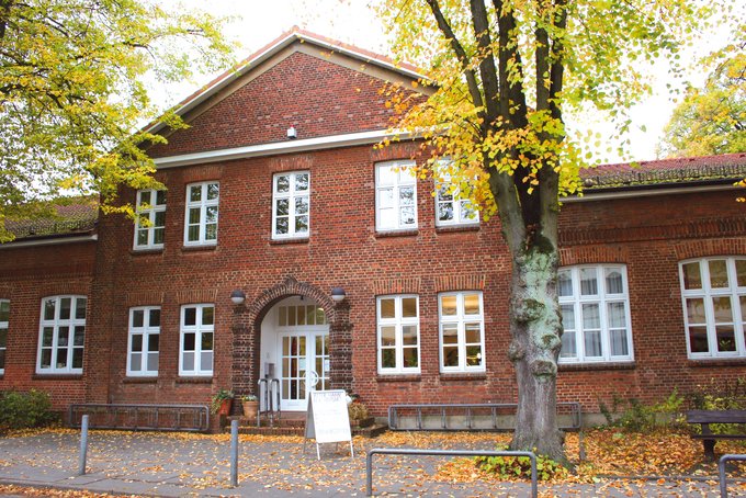 Backsteingebäude mit weißen Fenstern und Eingangstür, umgeben von Bäumen mit herbstlichem Laub