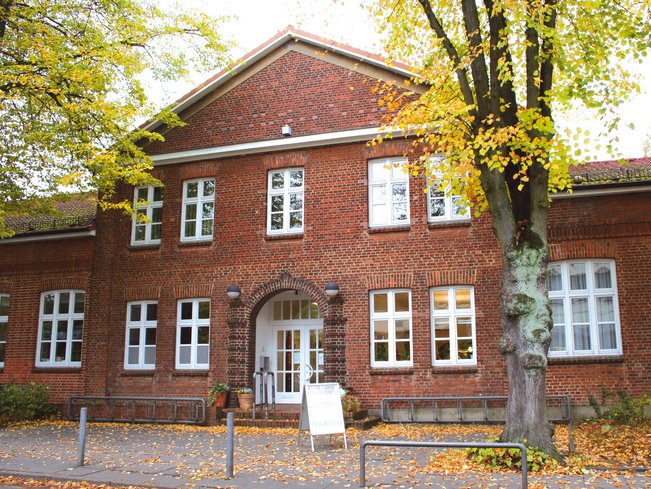 Backsteingebäude mit weißen Fenstern und Eingangstür, umgeben von Bäumen mit herbstlichem Laub