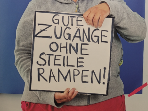 Person hält Schild auf dem steht "Gute Zugänge ohne steile Rampen"