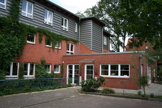 Mehrstöckiges Gebäude mit roten Ziegeln und grauen Fassaden, umgeben von Bäumen und einem kleinen Hof