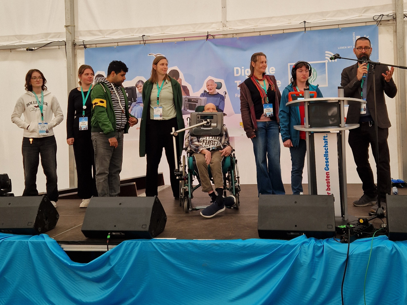 Sieben Personen auf einer Bühne bei einer Veranstaltung zur digitalen Teilhabe, darunter eine Person im Rollstuhl, eine Person spricht am Rednerpult mit Mikrofon.