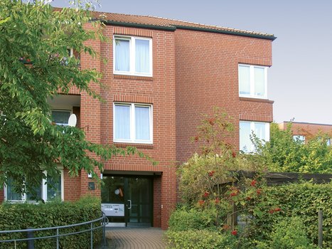 Mehrstöckiges Wohnhaus aus rotem Backstein mit weißen Fenstern, umgeben von Bäumen und Sträuchern