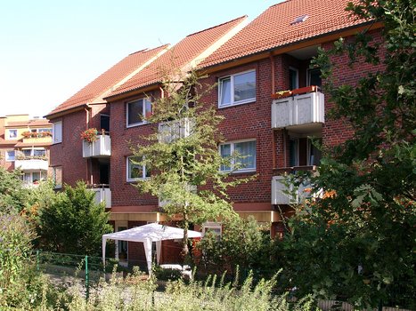 Außenansicht des Gebäudes. Mehrstöckiger roter Klinkerbau. Umgeben von einer Rasenfläche mit Büschen und Bäumen. Im Garten steht ein weißer Plastikpavillon zum Schutz vor Regen und Sonne. 