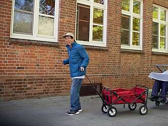 Person zieht roten Bollerwagen, begleitet von Rollstuhlfahrer und zwei weiteren Personen auf Gehweg vor Backsteingebäude