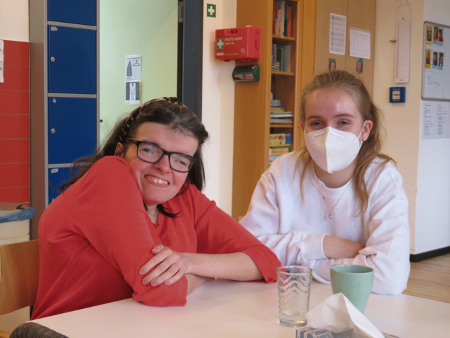 Mitarbeiterin mit Maske und Beschäftigte an einem Tisch