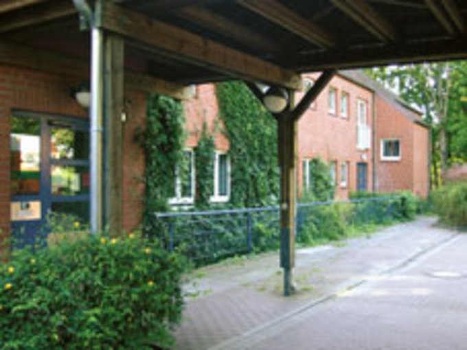 Überdachter Weg mit Holzstützen neben einem roten Backsteingebäude mit Fenstern und Efeu an der Fassade
