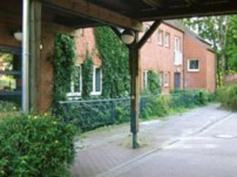 Überdachter Weg mit Holzstützen neben einem roten Backsteingebäude mit Fenstern und Efeu an der Fassade